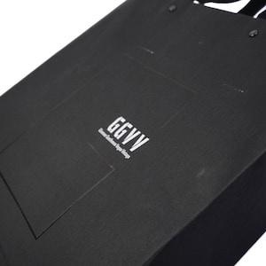 以下が含まれることがあります： 白文字で「GGVV」と書かれた黒いギフトバッグ。バッグは長方形で、表面はざらざらしています。「Glamour Gentlemen Vogue Vintage」の文字がロゴの下に印刷されています。