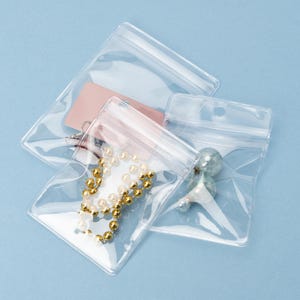 May include: Clear plastic zip-top bags containing various items. One bag holds a gold beaded necklace, another a pink tag, and a third contains a small, green, decorative object. The bags are arranged on a light blue surface.