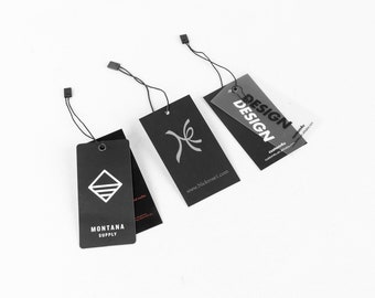 Pode incluir: Três etiquetas penduradas em preto e branco com um cordão amarrado. A primeira etiqueta tem um logotipo branco com o texto "MONTANA SUPPLY". A segunda etiqueta tem um logotipo branco com um "K" estilizado e o texto "www.Nickrosel.com". A terceira etiqueta é uma etiqueta de plástico transparente com o texto "DESIGN DESIGN" e "customdu" em branco.