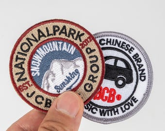 Może przedstawiać: Dwie haftowane naszywki. Jedna jest okrągła z tekstem "NATIONAL PARK" i "SNOW MOUNTAIN", grafiką gór i tekstem "JCB". Druga naszywka ma grafikę samochodu i tekst "CHINESE BRAND". Obie naszywki mają dodatkowy tekst.