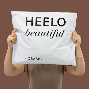 May include: White poly mailer with the words "HEELO beautiful" in black, with "JCBASIC" printed below. The mailer is held up by a person against a brown background.