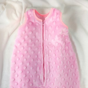 May include: A soft, pink baby sleep sack with a textured dot pattern. The sleeveless sack has a full-length zipper down the front and a pink trim around the neck and armholes. The sleep sack is designed to keep babies warm and secure.