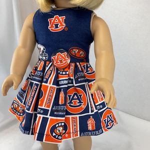 May include: A doll wearing a navy blue and orange dress with the Auburn University logo and text. The sleeveless dress has a fitted bodice and a flared skirt. The dress is adorned with the letter "A", the word "Tigers", and other Auburn University imagery.