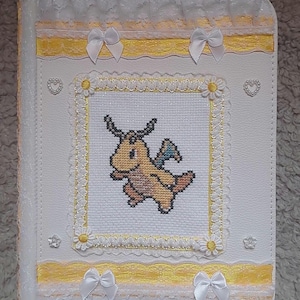 May include: White, handmade book cover with a cross-stitched design of a yellow dragon. The cover is adorned with yellow ribbon, white lace, and small white bows. Decorative hearts and flowers are also present.