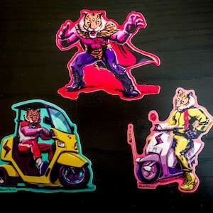 May include: Three colorful stickers featuring tiger-headed characters. One sticker depicts a tiger-headed figure in a superhero pose with a red cape. Another shows a tiger driving a yellow scooter. The third sticker shows a tiger driving a yellow three-wheeled vehicle.