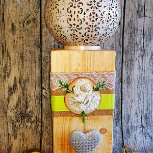 May include: A wooden candle holder with a white, decorative lantern on top. The holder is decorated with burlap ribbon, lace, a paper flower, and a silver glitter heart.