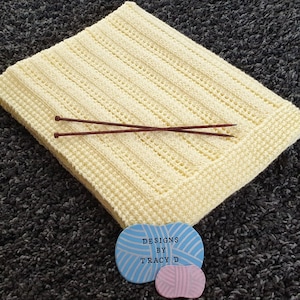 May include: A pale yellow knitted baby blanket with a simple stitch pattern. Two wooden knitting needles are lying on top of the blanket. A blue and white circular tag with the text "Designs by Tracy D" is in the lower right corner of the image.