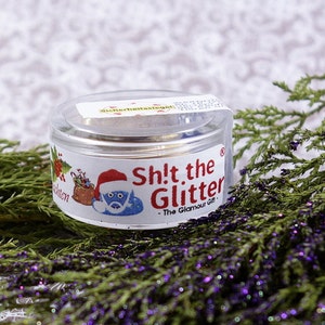 May include: A small, clear jar with a white label that says "Sh!t the Glitter - The Glamour Gift". The label also features a cartoon Santa Claus with a blue beard. The jar is sitting on a bed of green and purple foliage.