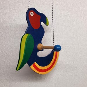 May include: A brightly coloured wooden parrot toy. The parrot has a blue body, red head, and green and yellow wings. It hangs from two black and white strings. A wooden dowel with a blue ball is attached.