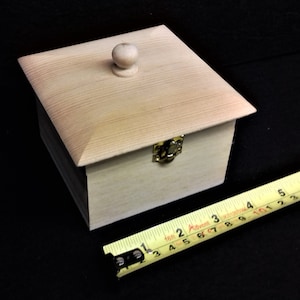 May include: A small wooden box with a hinged lid and a knob on top. The box is made of light wood and has a brass latch. A yellow measuring tape is in the foreground.