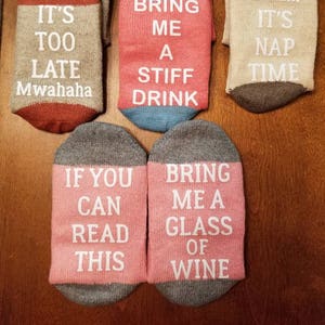 Op de afbeelding: Zes paar kleurrijke sokken met humoristische spreuken. De sokken zijn gemaakt van een zacht, comfortabel materiaal en zijn verkrijgbaar in verschillende kleuren, waaronder rood, blauw, grijs en roze. De spreuken op de sokken zijn: "Als je kunt, is het te laat Mwahaha", "Als je me een sterke drank brengt", "Sst... Het is tijd voor een dutje", "Als je dit kunt lezen", en "Breng me een glas wijn".