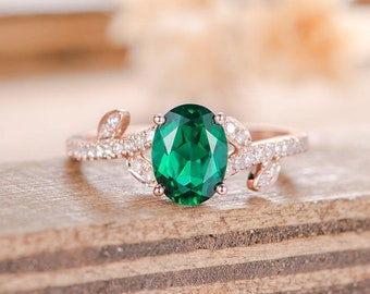 May include: A rose gold ring with a large oval emerald gemstone surrounded by smaller diamonds. The ring features a delicate leaf design on each side of the center stone.