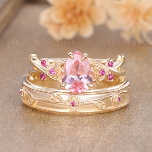 Peut inclure: Un ensemble de bagues dorées avec une pierre rose en forme de poire et de petites pierres roses. Les bagues présentent un motif floral avec des détails de roses. La marque "AUSGEMSCO" est visible en arrière-plan.