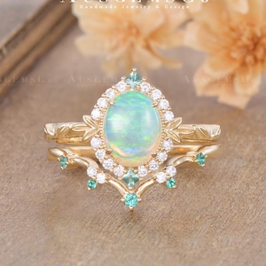 Peut inclure: Une bague de fiançailles dorée avec une pierre centrale en opale ovale, entourée d'un halo de petits diamants blancs. La bague présente de fins détails floraux et de petites pierres vertes. La bague est posée sur un fond neutre et doux.