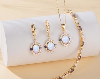 May include: A gold jewelry set featuring a necklace, earrings, and bracelet. Each piece has an oval opal center stone, surrounded by small clear and dark blue gemstones. The jewelry is displayed on a white surface with the text "AUSGEMSCO Handmade jewelry & Design".