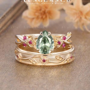 May include: A gold-colored ring set featuring a pear-shaped green gemstone and small red gemstones. The rings have floral and leaf details. The top ring has a curved design with the green gemstone at the center. The bottom ring has a floral and leaf pattern.