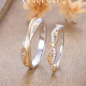 May include: Two wedding rings with a gold and silver design. One ring has a gold floral pattern and the other has a gold floral pattern with small diamonds. The rings are on a light brown surface. The text "AUSGEMSCO Handmade Jewelry & Design" is at the top.