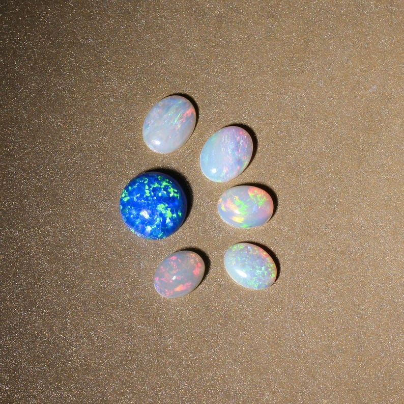 May include: A collection of seven oval-shaped opals, six white and one blue with green and white flecks, arranged in a semi-circle on a brown surface.