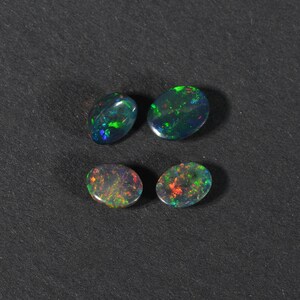 May include: Four oval-shaped black opals with green, red, and blue iridescent flashes. The opals are arranged in two rows of two on a black surface.