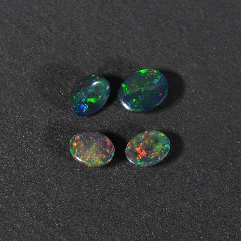 May include: Four oval-shaped black opals with green, red, and blue iridescent flashes. The opals are arranged in two rows of two on a black surface.