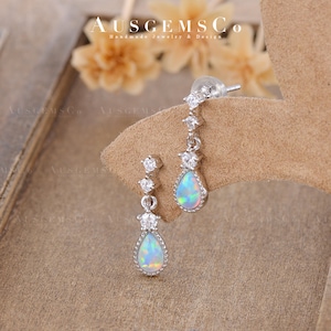 May include: A pair of silver teardrop earrings with opal stones and small clear cubic zirconia stones. The earrings are on a brown suede background.