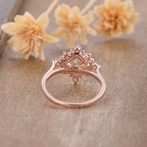 K&ouml;nnte beinhalten: Ein Verlobungsring aus Ros&eacute;gold mit einem quadratischen Mittelst&uuml;ck und einem Heiligenschein aus kleineren Steinen. Der Ring liegt auf einer Holzoberfl&auml;che.