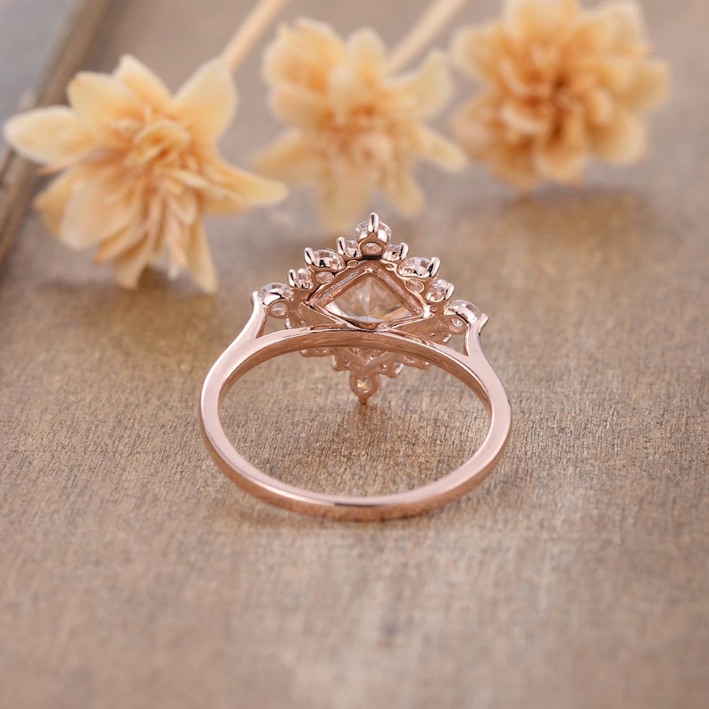 K&ouml;nnte beinhalten: Ein Verlobungsring aus Ros&eacute;gold mit einem quadratischen Mittelst&uuml;ck und einem Heiligenschein aus kleineren Steinen. Der Ring liegt auf einer Holzoberfl&auml;che.