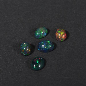 May include: Five oval-shaped opals with iridescent colours, including green, blue, and red, on a black background.