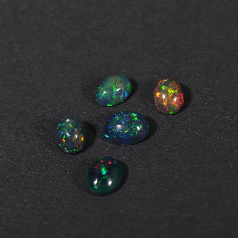 May include: Five oval-shaped opals with iridescent colours, including green, blue, and red, on a black background.