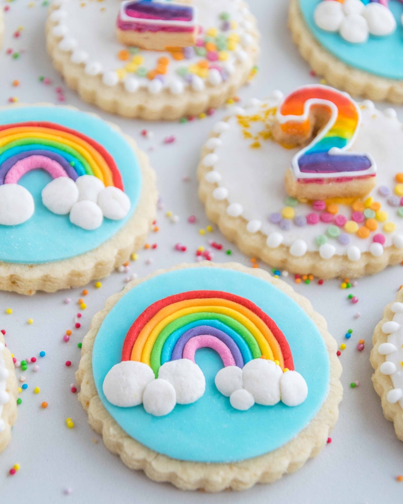 Op de afbeelding: Een close-up van regenboogversierde suikerkoekjes. De koekjes zijn versierd met kleurrijk glazuur en sprinkles. E&eacute;n koekje heeft een regenboog met wolken, een ander heeft een nummer twee en een ander heeft een regenboog met wolken.