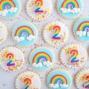 Op de afbeelding: Een dozijn ronde suikerkoekjes versierd met blauwe glazuur, regenboogsprankels en een nummer twee. Sommige koekjes hebben een regenboog met wolken erop, terwijl andere een nummer twee met regenboogsprankels hebben.