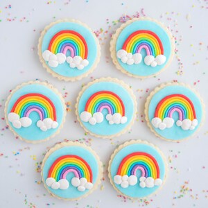 Op de afbeelding: Zeven ronde suikerkoekjes versierd met blauwe glazuur en regenboog fondant. Elk koekje heeft een regenboog met witte wolken.