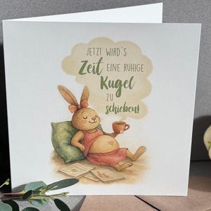 Op de afbeelding: Een wenskaart met een aquarel illustratie van een ontspannen konijn met een kopje koffie. Het konijn draagt een rode jurk en rust op een groen kussen. De kaart heeft tekst in een tekstballon.