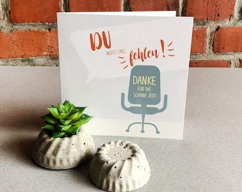 May include: A white card with the text "DU WIRST UNS FEHLEN!" in red and orange. The card also has a gray office chair with the text "DANKE FUR DIE SCHONE ZEIT" in yellow. The card is sitting on a gray surface with two small concrete planters with green succulents.
