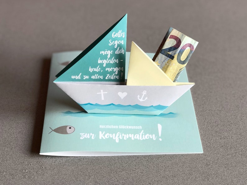 May include: A light blue card with a white paper sailboat on top. The sailboat has a yellow sail and a twenty euro bill inside. The card has the text "Gottes Segen m&ouml;ge dich begleiten - heute, morgen und zu allen Zeiten!" and "Herzlichen Gl&uuml;ckwunsch zur Konfirmation!" on it.