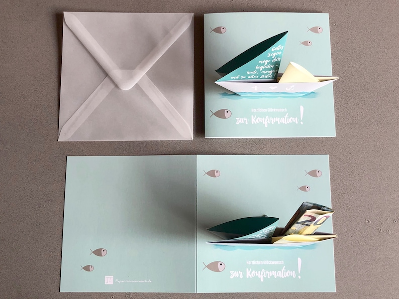 May include: A light blue pop-up card with a sailboat design. The sailboat is made of paper and has a green sail. The card has the text "zur Konfirmation!" on the front. The card is open and the sailboat is popped up. There are small fish on the front and inside of the card.