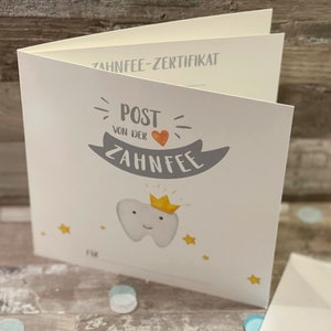 Może przedstawiać: Biała karta z wzorem certyfikatu wr&oacute;żki zębowej. Karta przedstawia kresk&oacute;wkową zębatkę z koroną, sercem i tekstem "Post von der Zahnfee".