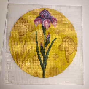 May include: A needlepoint design featuring a purple and white iris flower with green leaves and buds, set against a yellow circular background. The background includes faint floral outlines. The design is on a white mesh canvas.