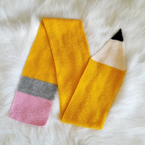 May include: A knitted scarf designed to resemble a pencil. The scarf is primarily yellow, with a gray and pink band at one end, and a white and black pencil tip at the other. The scarf is made of soft yarn.