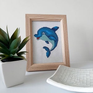 May include: A framed cross-stitch embroidery of a blue dolphin with a white background. The dolphin is smiling and has a red mouth.