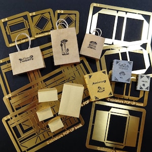 Może przedstawiać: Zestaw złotych metalowych szablonów do tworzenia miniaturowych książek pop-up. Szablony zawierają projekty książki z ilustracją grzyba, książki z tekstem "Kocham grzyby", książki z tekstem "Najlepsze życzenia", książki z tekstem "Rocznica" oraz książki o prostym wzorze. Szablony są oznaczone literami i cyframi.