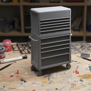 Puede incluir: Un cofre de herramientas gris con múltiples cajones y un compartimento superior. El cofre tiene un asa a cada lado y se asienta sobre cuatro pequeñas ruedas. El cofre de herramientas está sobre una superficie de madera.