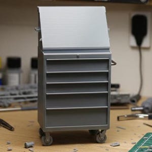 Op de afbeelding: Een grijze metalen gereedschapskist met een scharnierend deksel en meerdere laden. De gereedschapskist staat op wielen en heeft een handvat aan de zijkant. De gereedschapskist staat op een houten oppervlak.