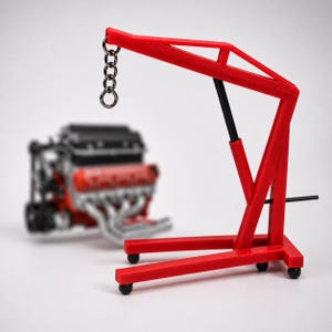 Peut inclure: Un modèle rouge de grue de moteur avec une chaîne et des roues noires. La grue est devant un modèle de moteur rouge et noir flou. La grue a une tige de support noire et une barre horizontale.