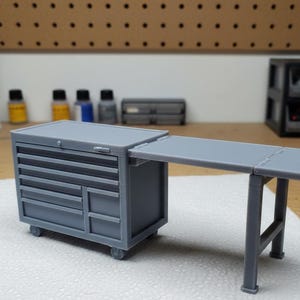 May include: A grey miniature tool chest with a fold-out work surface. The chest has multiple drawers and a small cabinet door. The work surface is supported by two legs. The tool chest is on wheels.