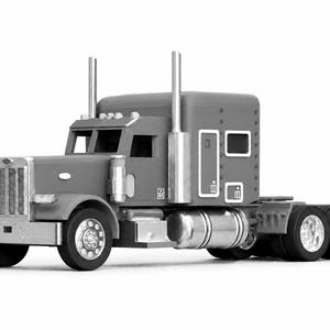 May include: A grey semi-truck model featuring a prominent front grille, dual vertical exhaust stacks, and a sleeper cab. The vehicle includes detailed silver-coloured fuel tanks, black tyres with metallic rims, and a rectangular chassis design.