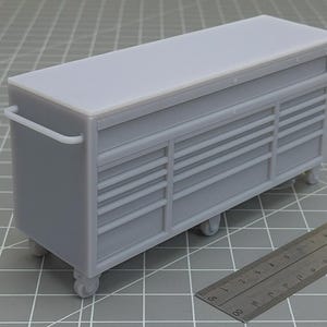 May include: A light grey miniature tool chest with multiple drawers and a flat top. The chest has a handle on one side and sits on small wheels. A ruler is visible, suggesting a scale model or tool organiser.