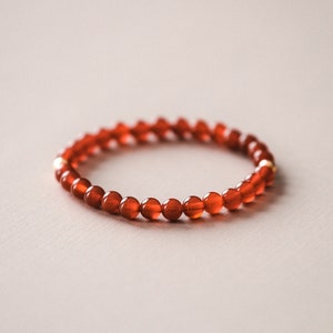 May include: A bracelet made of small, round, translucent orange-red beads. The beads are strung together, with a few gold-coloured beads. The jewellery is displayed on a neutral background.
