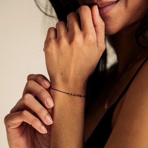 May include: A close-up of a person's wrist wearing a delicate black string bracelet with small, colorful beads.