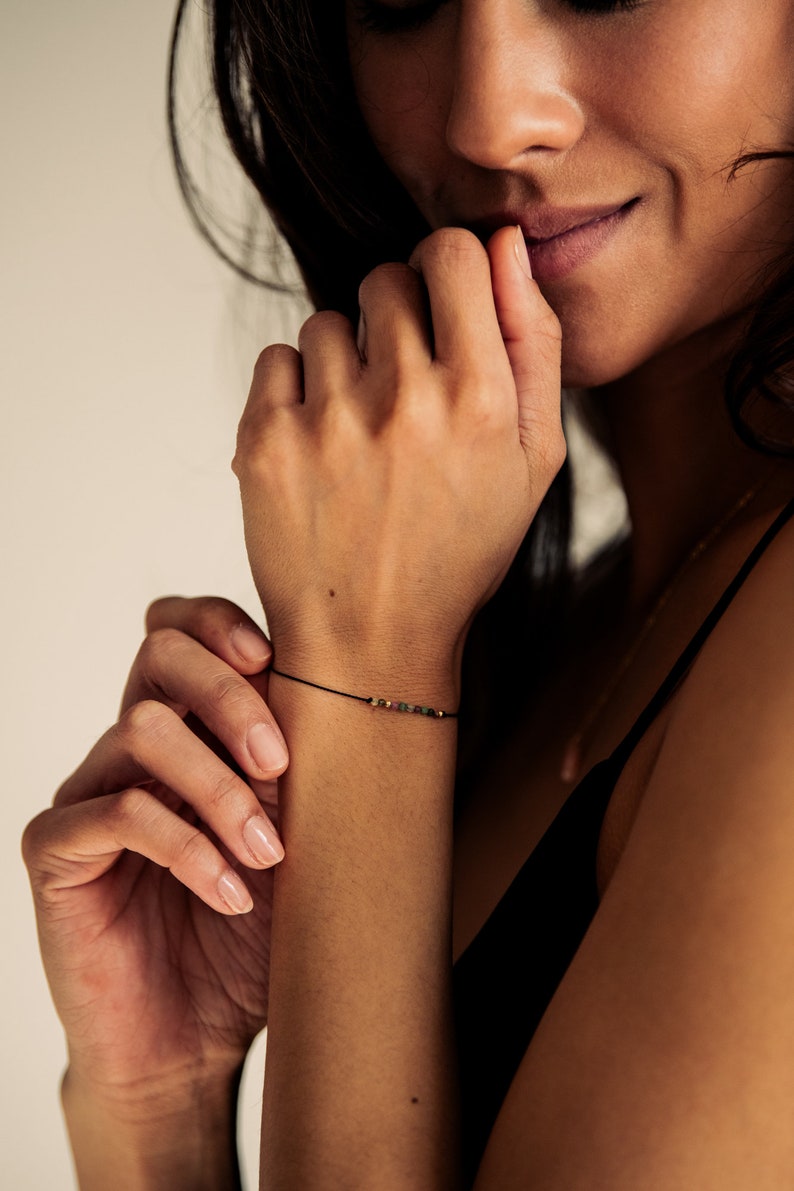 May include: A close-up of a person's wrist wearing a delicate black string bracelet with small, colorful beads.
